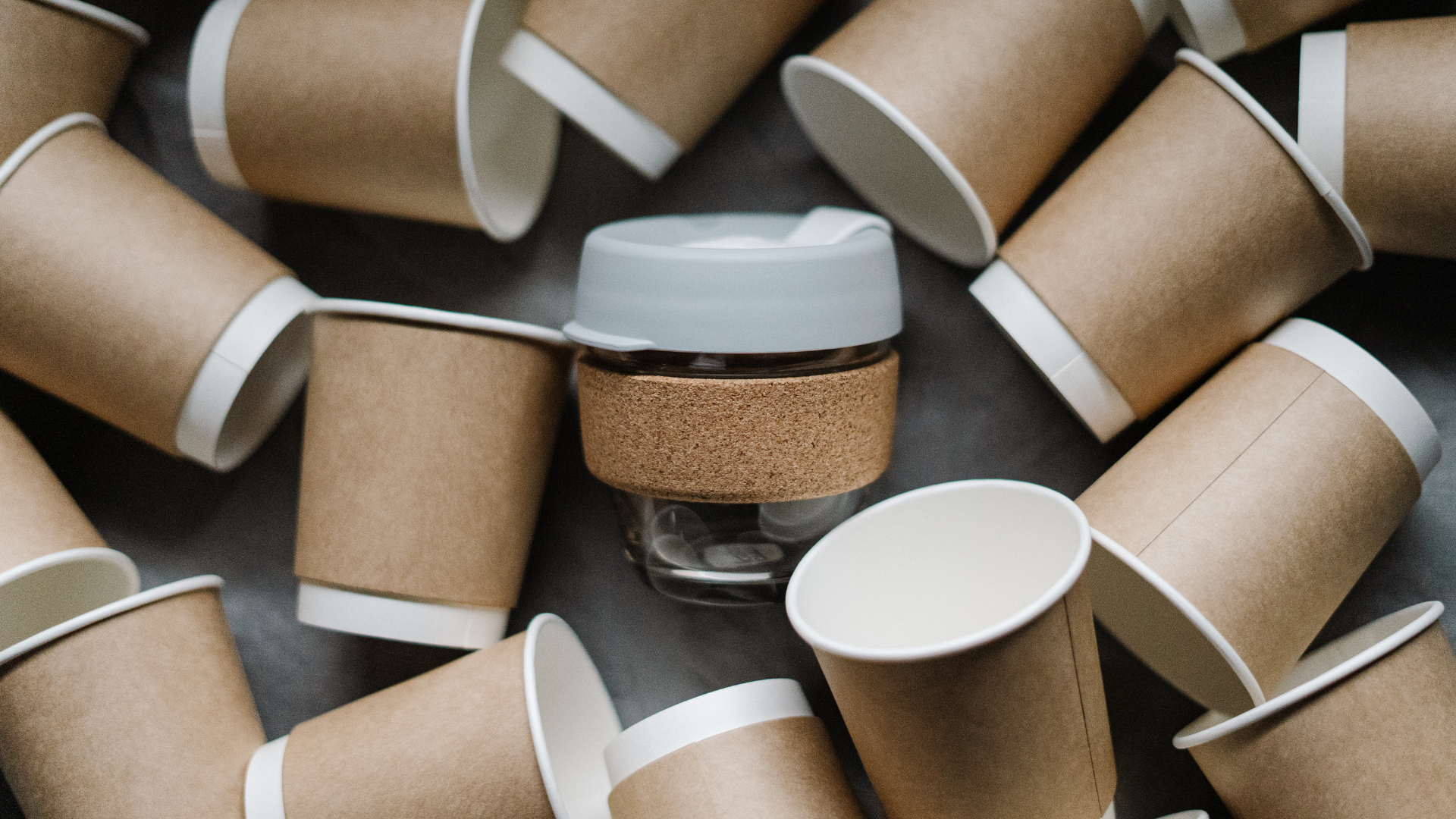 A group of brown paper cups scattered on a surface with a reusable glass cup featuring a cork sleeve in the center.