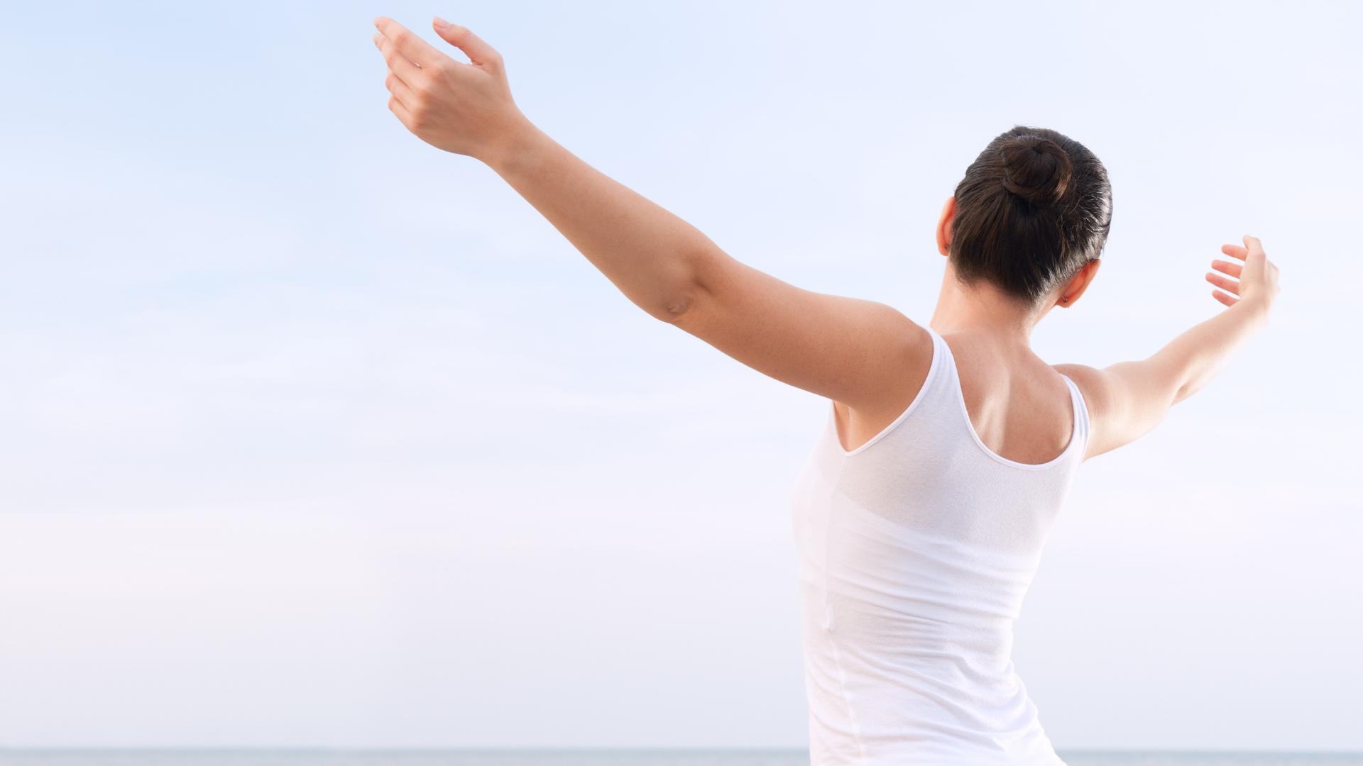 Woman in a white tank top standing by the sea with arms outstretched, symbolizing health, vitality, and wellness.