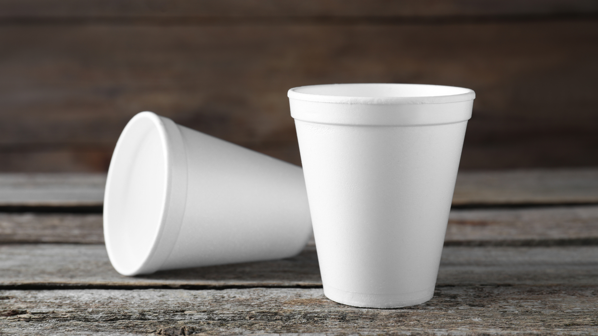 Two white polystyrene foam coffee cups on a rustic wooden surface, one standing upright and the other lying on its side.